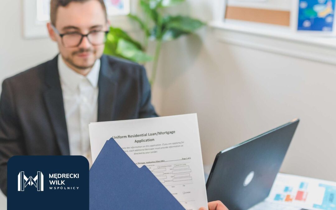 Mężczyzna w garniturze siedzący przy biurku, w tle roślina doniczkowa. Na pierwszym planie formularz dotyczący wniosku hipotecznego. Logo Kancelarii Mędrecki Wilk i Wspólnicy.