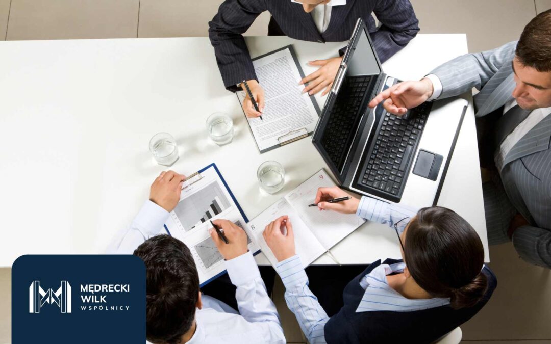 Grupa osób w biznesowym otoczeniu omawiająca dokumenty i analizy finansowe przy stole. Na pierwszym planie laptop i raporty. W lewym dolnym rogu logo kancelarii Mędrecki Wilk i Wspólnicy.
